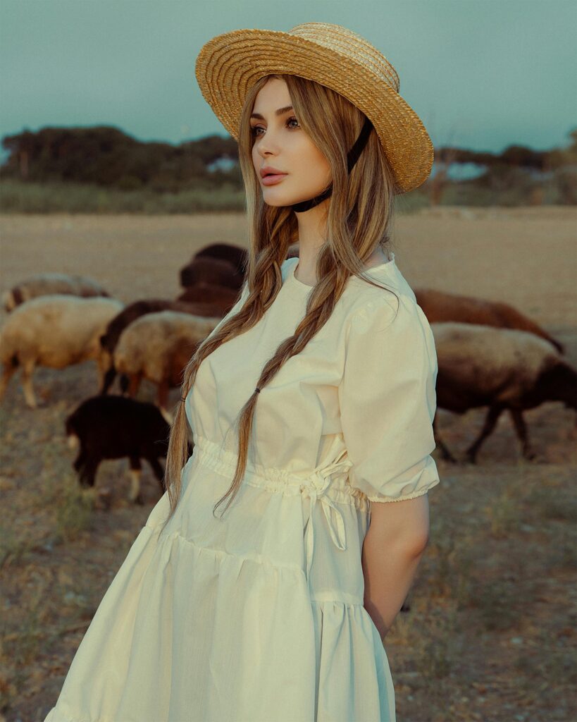 A woman in a white dress and straw hat stands in a field with sheep.